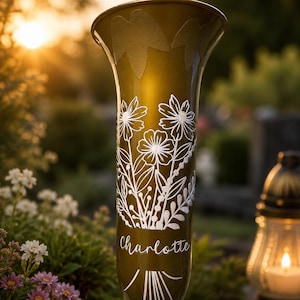 Könnte beinhalten: Eine trompetenförmige Grabvase in Metallic-Gold mit einer weißen Linienzeichnung eines Wildblumenstraußes und dem in Schreibschrift geschriebenen Namen Charlotte. Die Vase hat einen spitz zulaufenden Fuß und einen breiten, ausgestellten Rand.