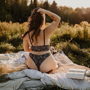 May include: A black lace lingerie set, comprising a bra and high-waisted bottoms, worn by a person sitting on a blanket in a field. The lingerie has a floral pattern. A book and flowers are also visible in the sunlit scene.