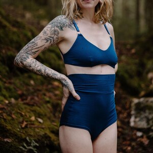 May include: A two-piece set of navy blue lingerie. The top is a triangle-style bra with thin straps. The high-waisted bottoms have a wide waistband. The woman in the image has tattoos on her arm and is standing in a forest.