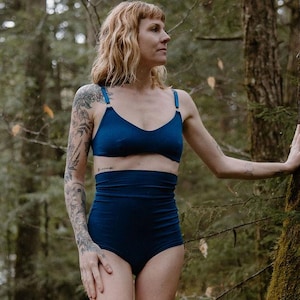 May include: A two-piece navy blue lingerie set. The top is a triangle-style bra with adjustable straps. The high-waisted bottoms offer full coverage. The model is standing in a forest setting.