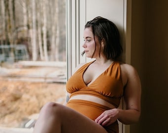 May include: A two-piece, mustard-colored swimsuit. The top is a halter style, and the bottoms are high-waisted. The person is sitting on a window sill, looking out at a blurred outdoor scene. The room has neutral-colored walls.