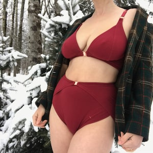 Op de afbeelding: Een bordeauxrode lingerie set, bestaande uit een beha en een slip met hoge taille, met goudkleurige sluitingen. De beha heeft verstelbare bandjes en een sluiting aan de voorkant. De slip heeft een hoge taille. Een groen-bruin geruit jasje wordt gedragen. Besneeuwd bos.