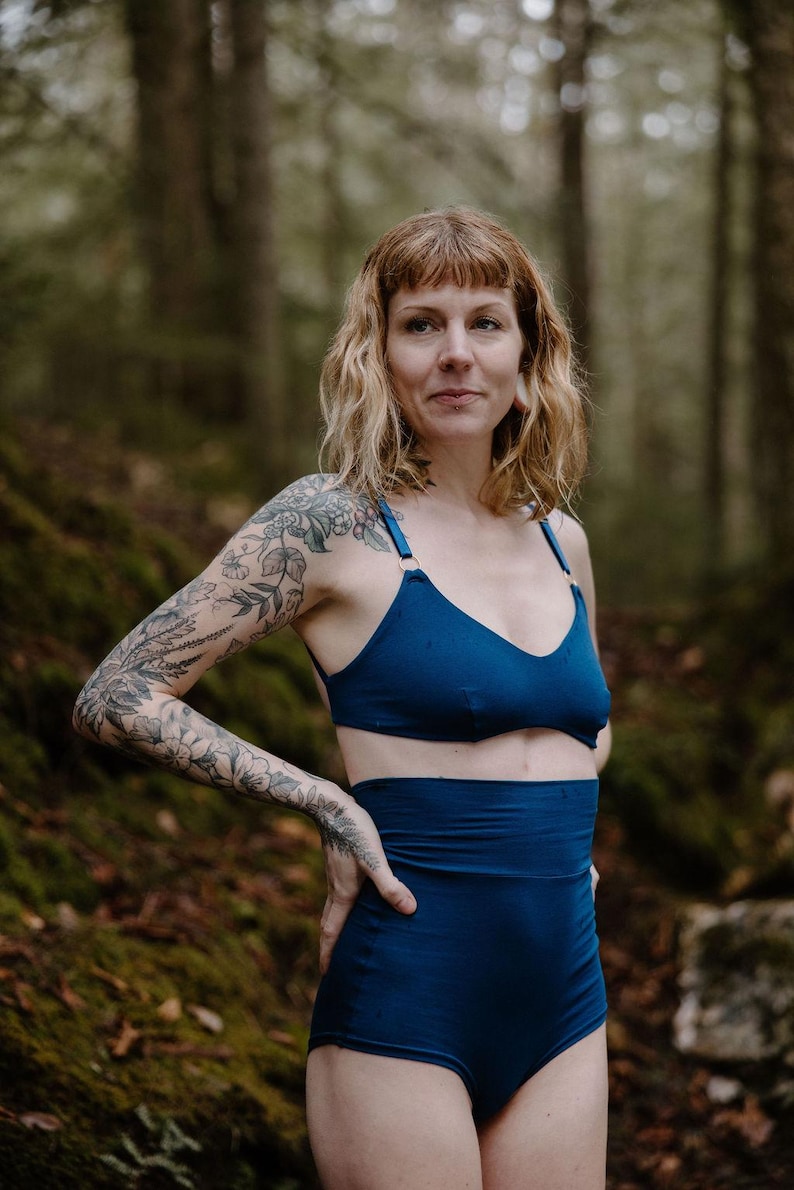 May include: A two-piece set of navy blue lingerie. The top is a triangle-style bra with thin straps. The high-waisted bottoms have a wide waistband. The woman in the image has tattoos on her arm and is standing in a forest.