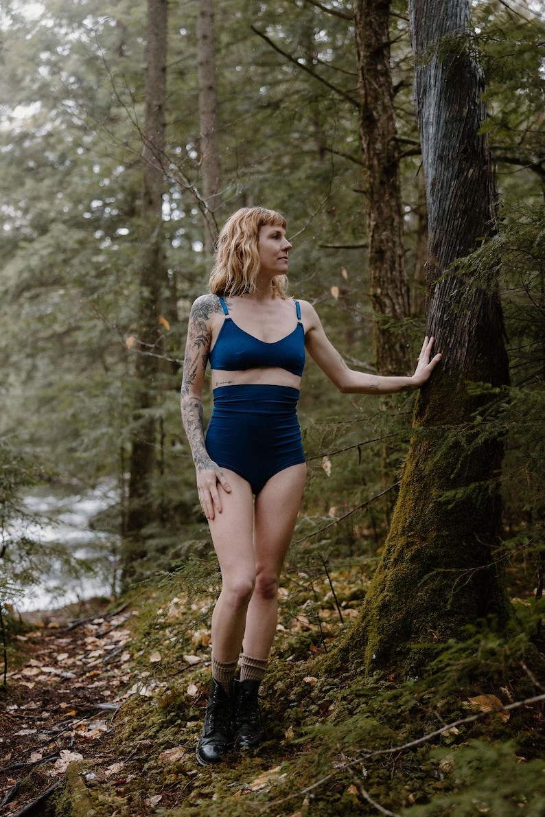 May include: A two-piece navy blue lingerie set. The top is a triangle-style bra with adjustable straps. The high-waisted bottoms offer full coverage. The model is standing in a forest setting.