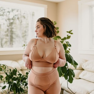 May include: A matching set of tan-colored lingerie. The set includes a triangle-style bra with adjustable straps and high-waisted shorts. The fabric appears to be a soft, knit material. The model is standing in a well-lit room with plants.