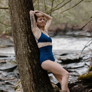 May include: A woman wearing a dark blue bikini set, consisting of a triangle top and high-waisted bottoms. The model is leaning against a tree trunk in a natural outdoor setting, with a river in the background.