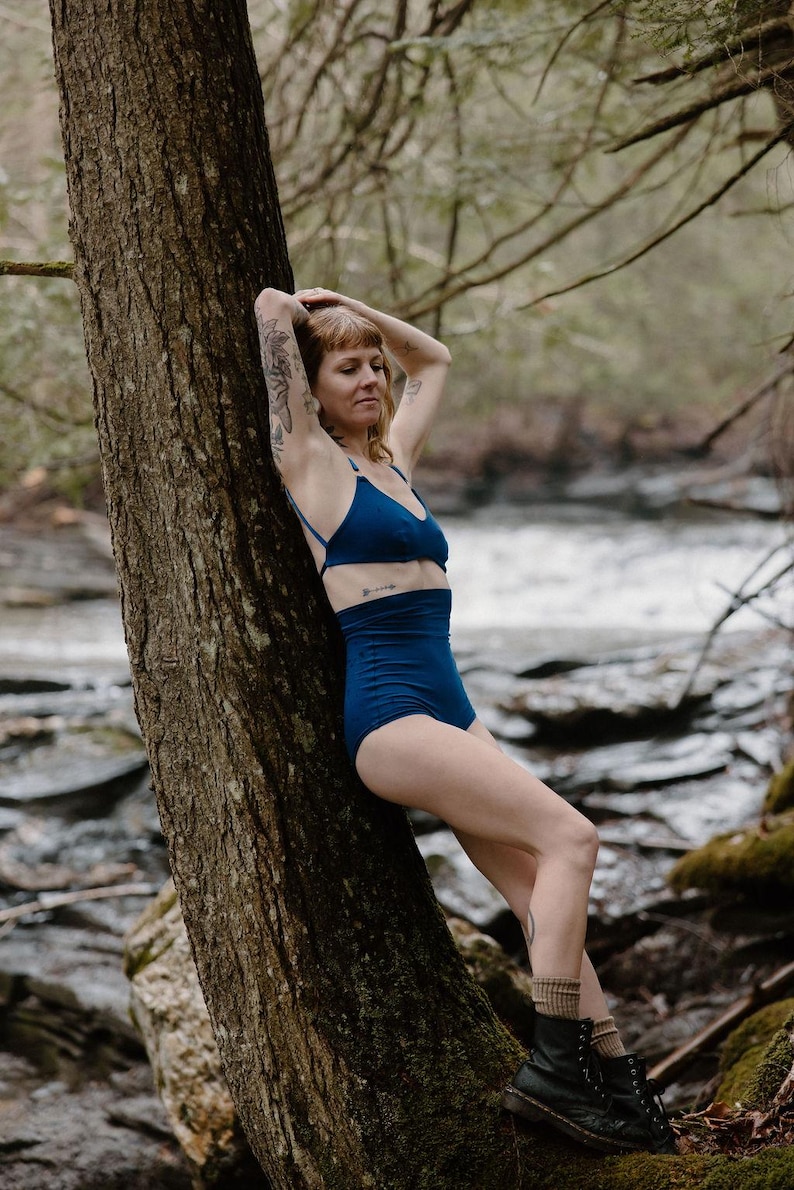 May include: A woman wearing a dark blue bikini set, consisting of a triangle top and high-waisted bottoms. The model is leaning against a tree trunk in a natural outdoor setting, with a river in the background.