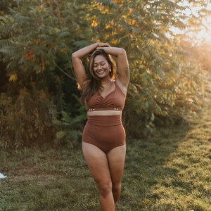 May include: A woman wearing a rust-colored bikini set, consisting of a wrap-style top and high-waisted bottoms. The bikini is made of a soft, solid-colored fabric. The woman is standing in a grassy area with trees in the background, lit by sunlight.