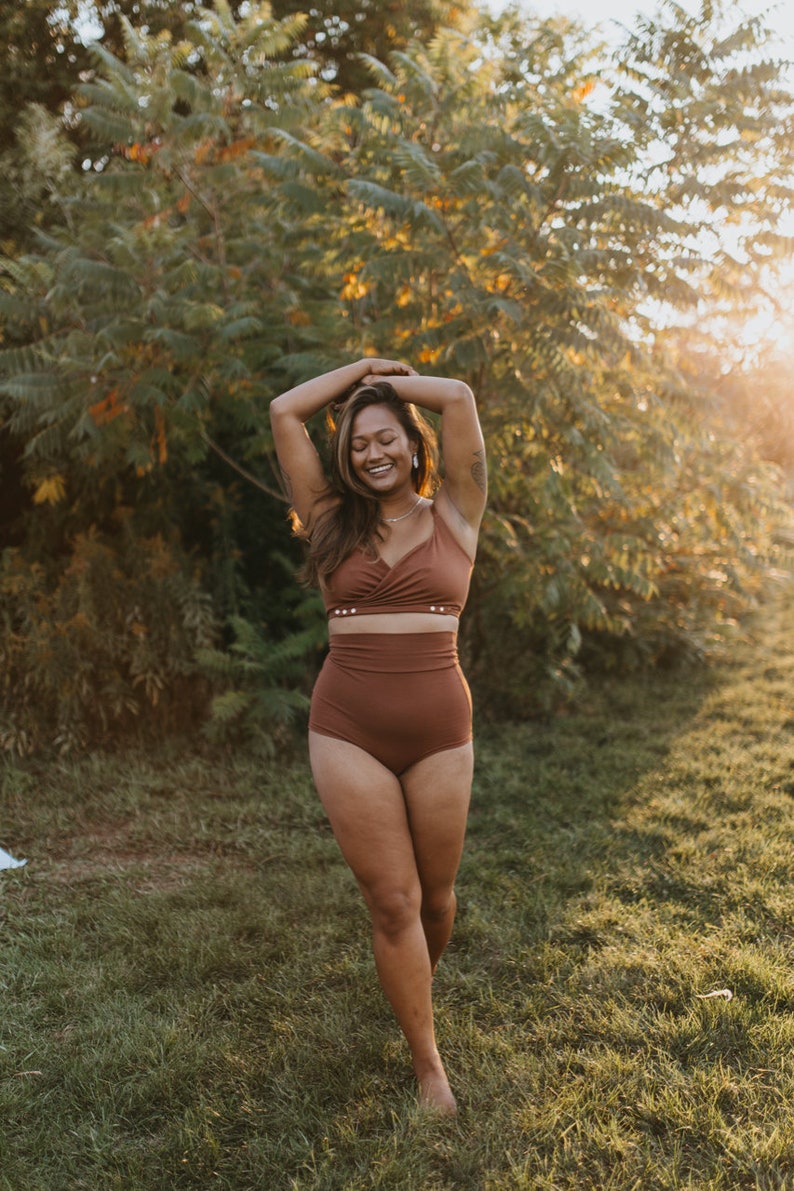 May include: A woman wearing a rust-colored bikini set, consisting of a wrap-style top and high-waisted bottoms. The bikini is made of a soft, solid-colored fabric. The woman is standing in a grassy area with trees in the background, lit by sunlight.