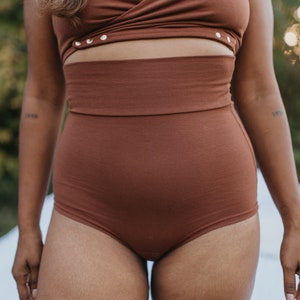 May include: A two-piece brown outfit. The top is a wrap-style design with gold-colored button details. The high-waisted bottoms are a solid brown color. The outfit is made of a soft, textured fabric. The model has a tattoo on their arm.