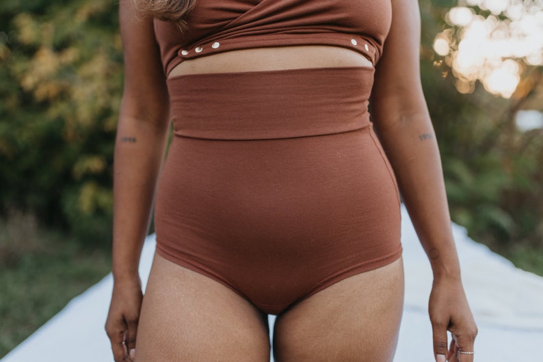 May include: A two-piece brown outfit. The top is a wrap-style design with gold-colored button details. The high-waisted bottoms are a solid brown color. The outfit is made of a soft, textured fabric. The model has a tattoo on their arm.