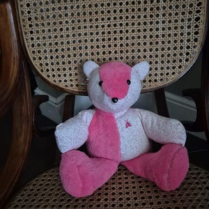 May include: A pink and white teddy bear with a floral pattern sits on a woven chair.