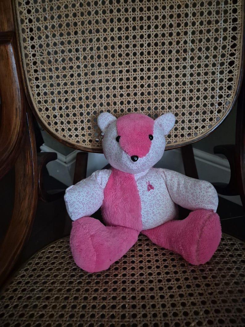 May include: A pink and white teddy bear with a floral pattern sits on a woven chair.