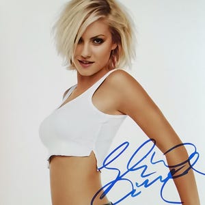 May include: A signed photograph featuring a woman with short blonde hair, wearing a white tank top and dark jeans. The autograph is in blue ink. The photo is on a white background.