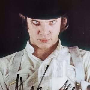 May include: A signed photograph featuring a person wearing a black bowler hat, white shirt, and suspenders. The person has striking eye makeup and a stern expression. The signature and the name "ALEX" are visible on the photograph.