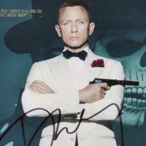 May include: Movie poster for "Spectre" featuring Daniel Craig in a white tuxedo holding a gun. The poster has a dark blue background with a skull image and the text "Spectre 007" and a signature.