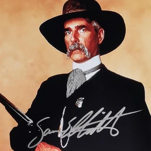 Könnte beinhalten: Farbfotografie eines Mannes in schwarzem Anzug, Hut und Krawatte, der ein Gewehr hält. Der Mann hat einen großen Schnurrbart und das Bild ist mit "Sam Elliott" signiert. Der Hintergrund ist ein Farbverlauf aus Orange und Beige.