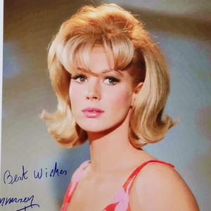 Op de afbeelding: Vintage foto van een vrouw met blond haar in een rode en roze bloemenjurk. De afbeelding bevat de handgeschreven tekst "Best Wishes" en een handtekening.