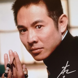 May include: A portrait of a man wearing a black outfit, holding a beaded necklace. The necklace features alternating black, brown, and cream-colored beads. The man's hand is in a prayer position. The image includes a signature in white.