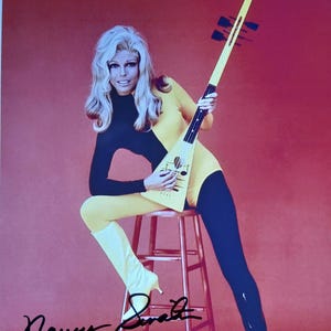 May include: A vintage photograph featuring a woman in a yellow and black jumpsuit, holding a unique, long-necked instrument. She is seated on a stool against a red background. The image is signed "Nancy Sinatra".