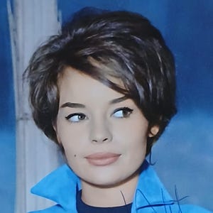 Puede incluir: Fotografía vintage de una mujer con cabello corto y oscuro, vestida con una chaqueta azul con cuello. La imagen tiene una estética retro, con una firma en la esquina inferior derecha. La mujer mira hacia un lado.