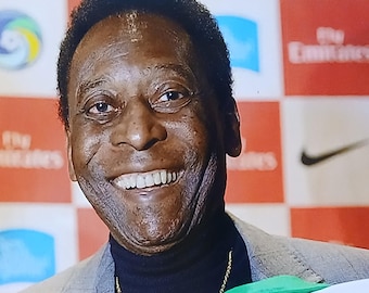 Może przedstawiać: Podpisane zdjęcie Pelé, brazylijskiej legendy piłki nożnej, uśmiechniętego. Trzyma zielono-białą koszulkę z jego imieniem dużymi zielonymi literami i podpisem. Tło przedstawia czerwono-biały wzór w szachownicę.