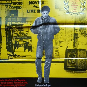 以下が含まれることがあります： 映画『タクシードライバー』の黄色と黒のポスター。ジャケットとジーンズ姿のロバート・デ・ニーロが街を歩く姿が描かれています。赤いタイトル、キャストとスタッフの名前、ドイツ語のテキストが入った月桂冠のグラフィックが含まれています。