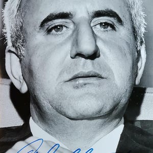 May include: Black and white portrait of a man in a suit and tie. The man has short, graying hair and a serious expression. A blue signature is visible at the bottom of the image.