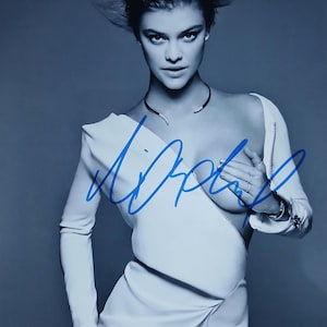 Peut inclure: Photographie en noir et blanc d'une femme portant une robe blanche avec un détail de fermeture éclair. La robe a des manches longues et un design asymétrique unique. Une signature est visible à l'encre bleue. La femme porte également un collier noir.