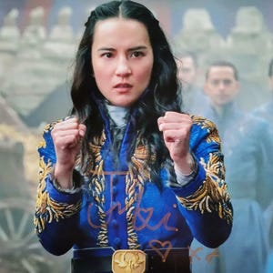 Könnte beinhalten: Eine Frau in einer blauen Jacke mit goldener Stickerei und einem schwarzen Gürtel mit goldener Schnalle. Sie ballt die Fäuste. Der Hintergrund zeigt verschwommene Figuren in blauen Mänteln und eine strohähnliche Struktur.