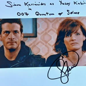 May include: A photograph of a man and a woman, with handwritten text above reading "Simon Kassianides as Yusef Kabira in 007 Quantum of Solace." The image is signed in black ink.