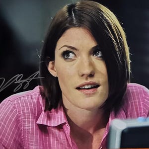 May include: A woman with dark hair and light eyes, wearing a pink collared shirt with a subtle striped pattern. The image includes a signature in the upper left corner. The background is dark and out of focus.
