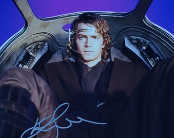 May include: A signed photograph featuring a person seated in a cockpit. The person is wearing a dark brown outfit and a headset. The background is a vibrant purple. The signature is in blue ink.