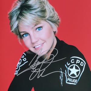 Puede incluir: Retrato de una mujer con pelo rubio corto y ojos azul claro, vestida con un uniforme negro con parches de "CPD POLICE". La imagen está firmada con tinta plateada. El fondo es rojo liso.