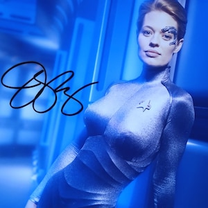 Può includere: Fotografia firmata con una donna in uniforme argentata di Starfleet. Ha un segno distintivo sul viso e un'insegna di Starfleet. Lo sfondo è un interno dai toni blu, e la firma è con inchiostro nero.