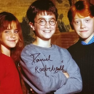 Puede incluir: Una foto grupal de tres jóvenes sonriendo a la cámara. La persona del centro lleva una sudadera gris con una firma manuscrita. La persona de la izquierda lleva una camiseta roja y la de la derecha un suéter negro.