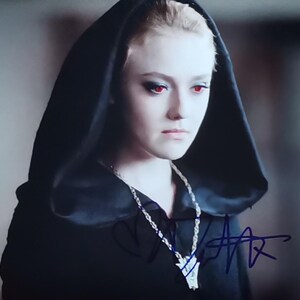 Puede incluir: Retrato de una persona con piel pálida y ojos rojos, vistiendo una capa con capucha negra. Se ve un collar plateado con un colgante. La imagen incluye una firma en la parte inferior.