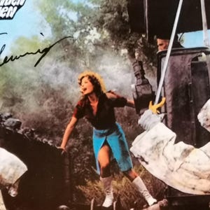 Może przedstawiać: Zdjęcie z filmu w stylu vintage przedstawiające kobietę w czerwonej koszuli i niebieskiej spódnicy, z mieczem i postaciami w białych szatach z kapturem. Obraz ma estetykę retro, z tekstem i podpisem w lewym górnym rogu.