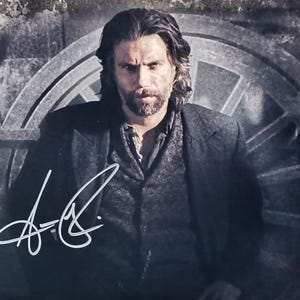 May include: A monochrome photograph of a man with long hair and a beard, wearing a dark coat and waistcoat. The image includes a signature in white ink. The background features a large, circular structure.