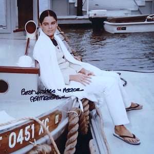 以下が含まれることがあります： 白いブレザーとパンツを含む白い服装を着た人がボートに座っています。その人はビーチサンダルを履いています。ボートには「0451 XC」の番号とロープが付いた木製のパネルがあります。画像には手書きのテキスト「Best wishes 4 Peace」が含まれています。