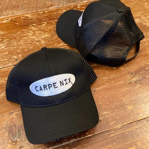 May include: Two black mesh trucker hats. One hat has a white oval patch with the text "CARPE NIX" embroidered on it.
