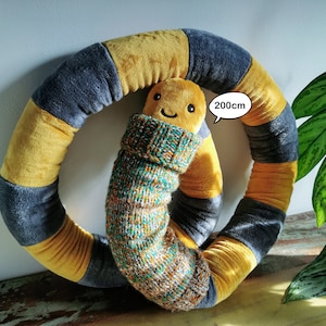 May include: A large, yellow and gray plush ring with a smaller plush worm in a green and brown knitted sweater. The worm is smiling and has a speech bubble that says "200cm".