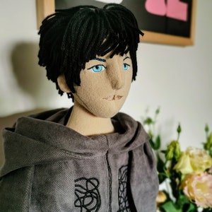 Puede incluir: Una muñeca de peluche con cabello negro, ojos azules y una sudadera con capucha gris. La muñeca tiene una expresión neutral y mira hacia la derecha.