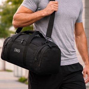Könnte beinhalten: Schwarze Sporttasche mit den Initialen "JMS" in Weiß gestickt. Die Tasche hat einen Schultergurt und mehrere Reißverschlussfächer. Die Person im Bild trägt ein graues T-Shirt und schwarze Shorts.