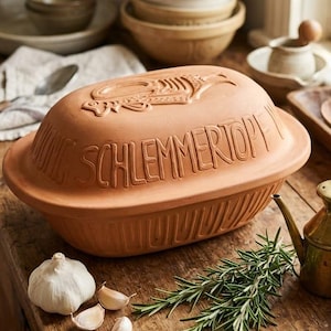 May include: An oval terracotta clay cooking pot with a lid featuring embossed text that reads SCHLEMMERTOPF and a stylized fish graphic. The unglazed earthenware vessel has a ribbed base and a natural reddish-brown finish.