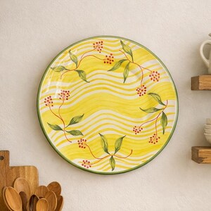 May include: A round ceramic plate featuring a vibrant yellow background with horizontal wavy white lines. The plate is decorated with a painted botanical pattern of thin brown stems, green leaves, and small clusters of red berries along the rim.
