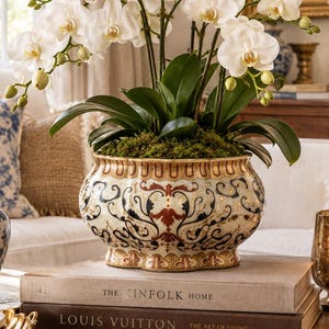 May include: A potted white orchid plant with multiple blooming flowers and green leaves sits in a decorative ceramic bowl featuring intricate gold, black, and rust-coloured patterns. The bowl rests on two stacked books titled The Kinfolk Home and Louis Vuitton.