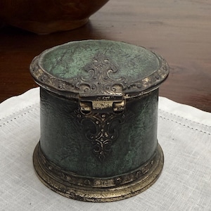 May include: A round, lidded box featuring a textured, dark green finish with distressed gold-toned metal accents. The lid and base are rimmed with a metallic border, and a decorative metal hinge plate with intricate scrollwork adorns the front.