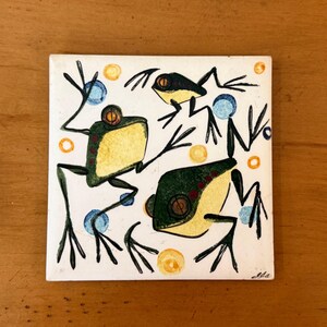 May include: A square ceramic tile featuring a white background with three illustrated frogs in shades of dark green and yellow. The design includes scattered blue and yellow circles, with black ink outlines defining the frogs and their limbs.