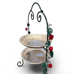 May include: A two-tiered serving stand with two aluminum foil pie pans. The dark green metal frame has decorative swirls and is adorned with red apple and green leaf accents. The stand is designed to hold food.